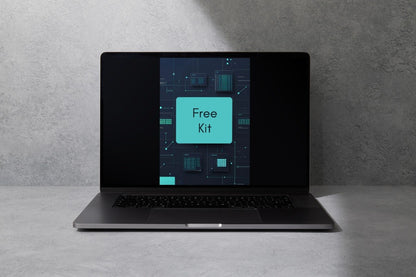 A dark gray laptop sits open on a light gray surface against a textured gray wall, with its screen displaying a digital graphic that reads 'Free Kit'.