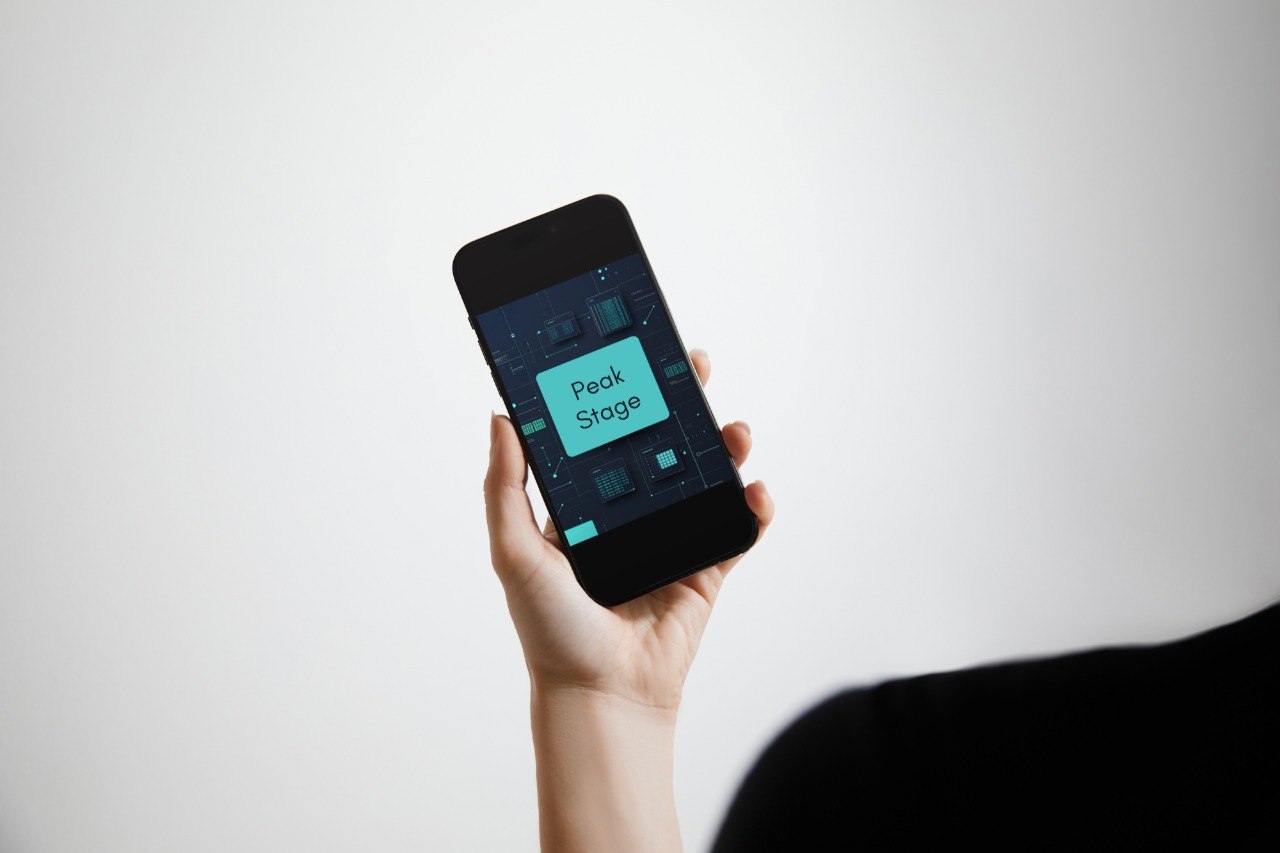A hand holding a black smartphone against a light gray background, with the phone screen displaying a dark interface and a bright teal label that reads 'Peak Stage'.