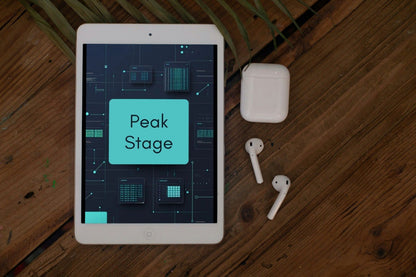 A white tablet lying on a wooden surface, displaying a digital interface with the words 'Peak Stage' in a teal box. To the right of the tablet are white wireless earbuds and their charging case.