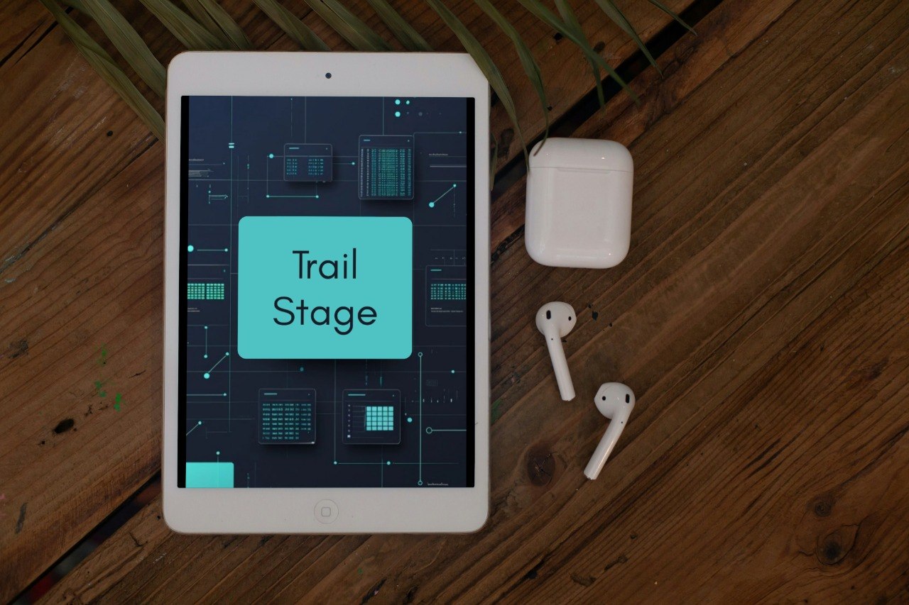 A tablet device displaying a digital interface labeled 'Trail Stage' lies on a wooden surface, alongside a white wireless earbud charging case and two white earbuds.